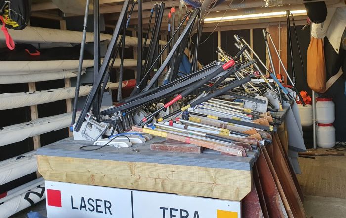 Image of sailing shed with masts rolled neatly down the left-hand side. Central storage area for rudders and daggerboards has been organised by dinghy type. There is one set of foils per slot.